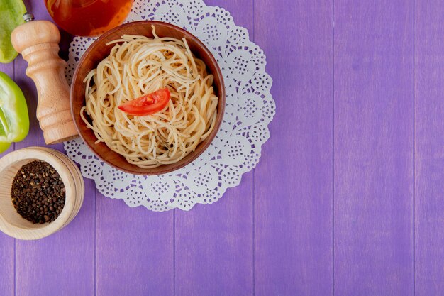 Draufsicht der Spaghetti-Nudeln in der Schüssel auf Papierdeckchen mit halb geschnittenem Pfefferbuttersalz und schwarzen Pfeffersamen auf lila Hintergrund mit Kopienraum