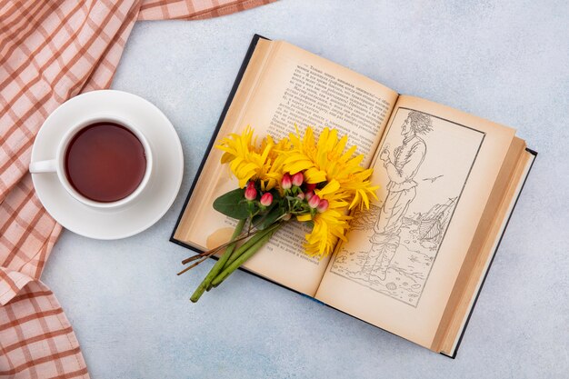 Draufsicht der Tasse Tee auf kariertem Stoff und Blumen auf offenem Buch auf Weiß