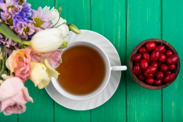 Draufsicht der Tasse Tee auf Untertasse und Schüssel Kornelkirschenbeeren mit Blumen auf grünem Hintergrund