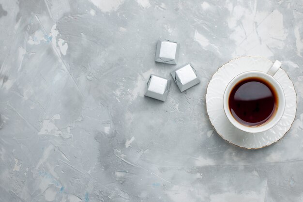 Draufsicht der Tasse Tee heiß in weißer Tasse auf Glasplatte mit Silberpaket Pralinen auf hellem Schreibtisch, Teegetränk süßer Schokoladenkeks