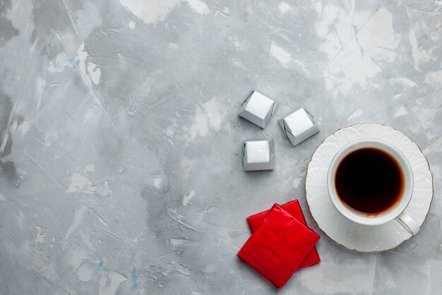 Draufsicht der Tasse Tee heiß in weißer Tasse auf Glasplatte mit Silberverpackung Pralinen auf hellem Schreibtisch, Teegetränk süße Schokoladenplätzchen Teatime