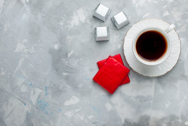 Draufsicht der Tasse Tee heiß in weißer Tasse auf Glasplatte mit Silberverpackung Pralinen auf weißem, Teegetränk süßer Schokoladenkeks