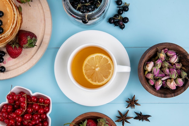 Draufsicht der Tasse Tee mit einer Zitronenscheibe mit roten und schwarzen Johannisbeeren und Erdbeeren auf einer blauen Oberfläche