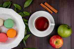 Kostenloses Foto draufsicht der tasse tee mit zimtfarbenen macarons und äpfeln auf einer holzoberfläche