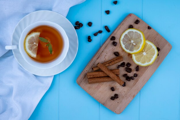 Draufsicht der Tasse Tee mit Zitronenscheibe auf weißem Stoff und Zimt mit Zitronenscheiben und Schokoladenstücken auf Schneidebrett auf blauem Hintergrund