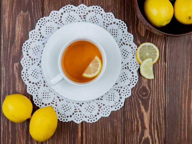 Draufsicht der Tasse Tee mit Zitronenscheibe darin auf Papierdeckchen und Zitronen auf hölzernem Hintergrund