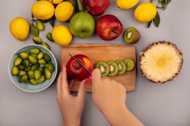 Kostenloses Foto draufsicht der weiblichen hände, die apfel auf einem hölzernen küchenbrett mit messer mit kinkans auf einer schüssel mit äpfeln kiwi-ananas und zitronen schneiden, die auf einer weißen wand lokalisiert schneiden