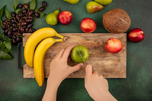 Draufsicht der weiblichen hände, die apfel mit messer und bananenpfirsich auf schneidebrett und birnen-kokosnuss-traubenpfirsich auf grünem hintergrund schneiden