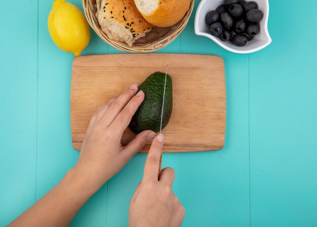 Draufsicht der weiblichen Hände, die Avocado mit einem Messer auf hölzernem Küchenbrett mit einem Eimer Brot mit schwarzen Oliven auf Schüssel auf Blau schneiden schneiden