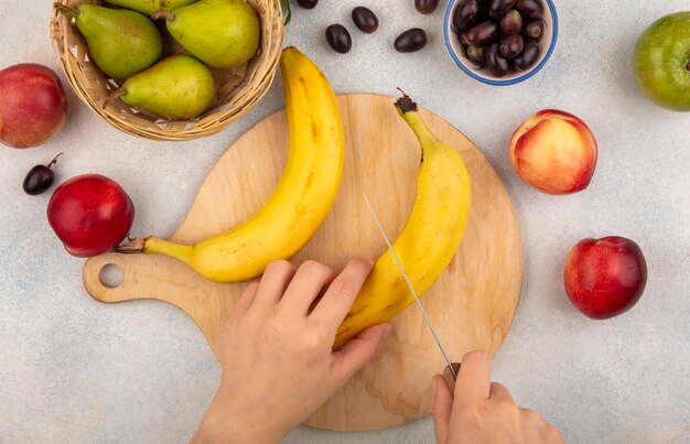 Draufsicht der weiblichen Hände, die Banane mit Messer auf Schneidebrett und Traubenbirnenpfirsichapfel auf weißem Hintergrund schneiden