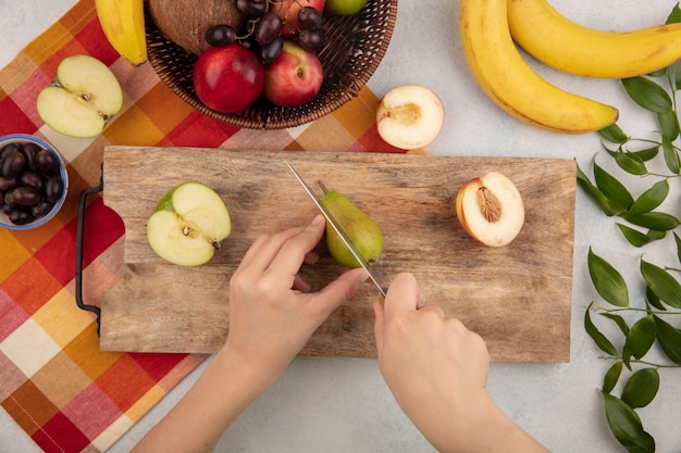 Draufsicht der weiblichen Hände, die Birne mit Messer und halbem Apfel und Pfirsich auf Schneidebrett und Korb der Pfirsich-Trauben-Kokosnuss auf kariertem Stoff mit Bananen und Blättern auf weißem Hintergrund schneiden