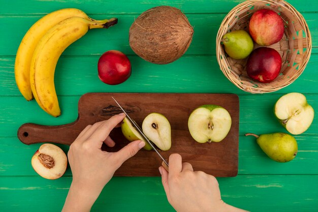 Draufsicht der weiblichen Hände, die Birne mit Messer und halben Apfel auf Schneidebrett mit Pfirsich-Bananen-Kokosnuss auf grünem Hintergrund schneiden