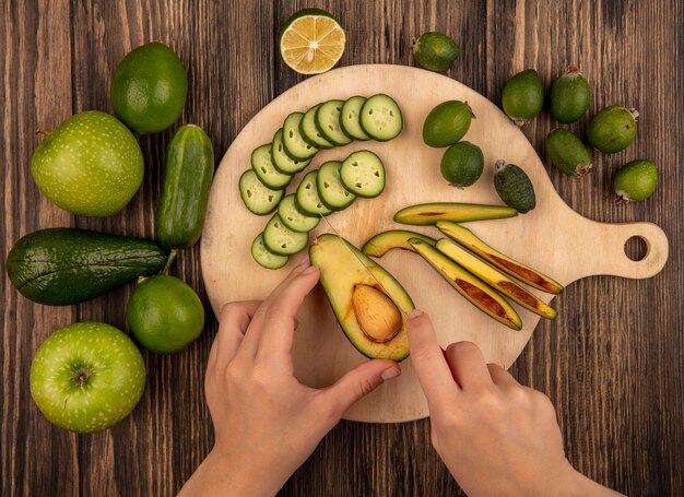 Draufsicht der weiblichen Hände, die eine Avocado mit Messer auf einem hölzernen Küchenbrett mit gehackten Gurkenscheiben mit den ganzen grünen Äpfeln der Gurkenlimetten und der Avocado schneiden, die auf einer hölzernen Oberfläche lokalisiert werden