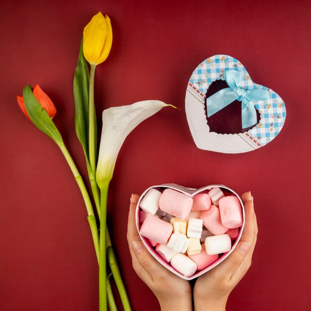 Kostenloses Foto draufsicht der weiblichen hände, die eine herzförmige geschenkbox halten, gefüllt mit marshmallow und roten und gelben farbtulpen mit callalilie auf rotem tisch