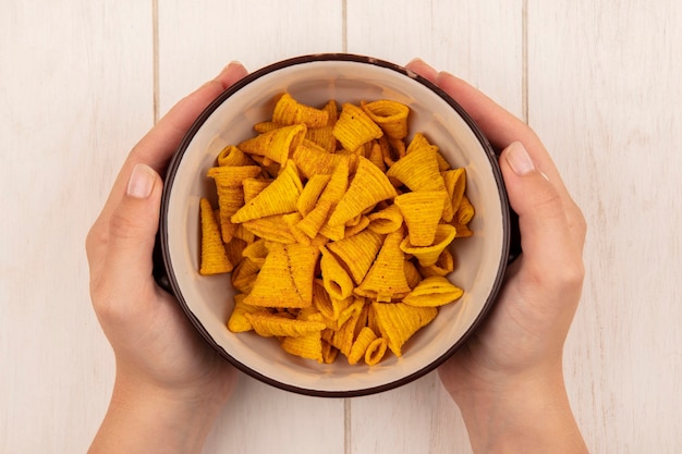 Draufsicht der weiblichen Hände, die eine Schüssel der knusprigen leckeren Chips auf einem beigen Holztisch halten