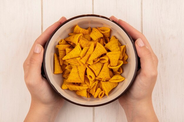 Draufsicht der weiblichen Hände, die eine Schüssel der knusprigen leckeren Chips auf einem beigen Holztisch halten