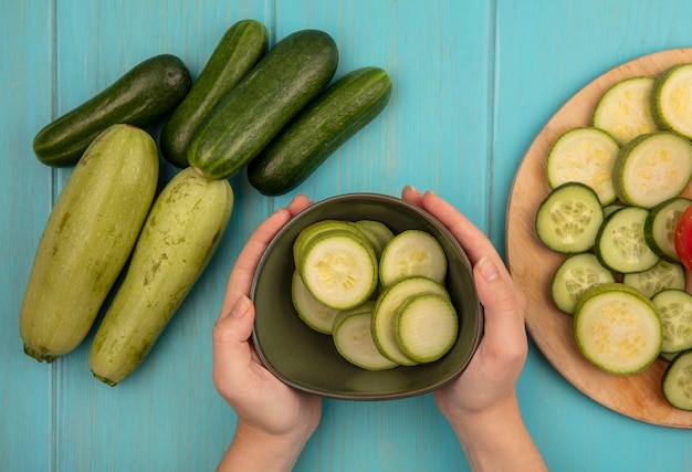 Draufsicht der weiblichen Hände, die eine Schüssel gehackte Zucchini mit Gurken und Zucchini halten, die auf einer blauen Holzoberfläche lokalisiert werden