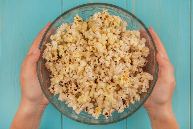 Draufsicht der weiblichen Hände, die eine Schüssel Popcorn auf einem blauen Holztisch halten