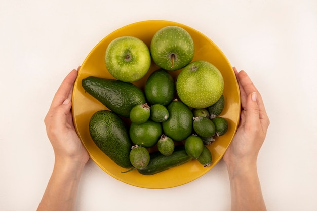 Draufsicht der weiblichen Hände, die einen gelben Teller mit frischen Früchten wie Äpfeln feijoas und Avocado auf einer weißen Oberfläche halten