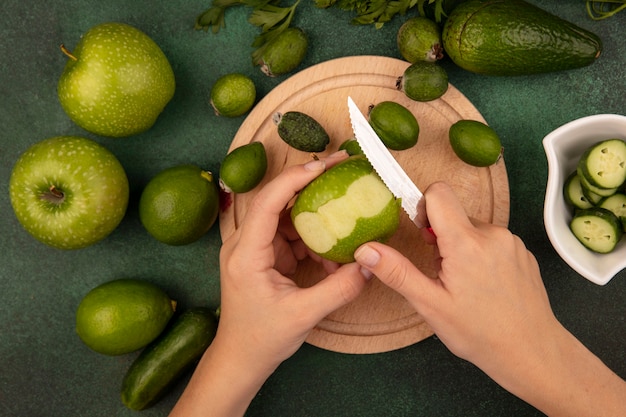 Draufsicht der weiblichen Hände, die einen grünen frischen Apfel mit Messer auf einem hölzernen Küchenbrett mit Limetten, Feijoas und grünen Äpfeln schälen, die auf einer grünen Oberfläche lokalisiert werden