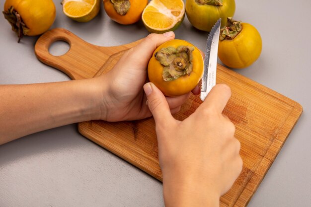Draufsicht der weiblichen Hände, die frische Kakifrucht auf einem hölzernen Küchenbrett mit Messer schneiden