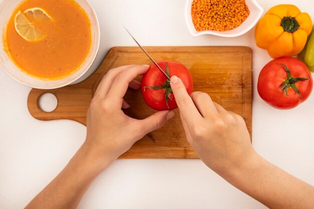 Draufsicht der weiblichen Hände, die frische Tomate auf einem hölzernen Küchenbrett mit Messer mit Linsensuppe auf einer Schüssel mit bunten Paprikaschoten lokalisiert auf einer weißen Oberfläche schneiden