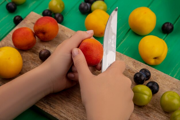 Draufsicht der weiblichen Hände, die frischen Pfirsich mit Messer auf einem hölzernen Küchenbrett auf einem grünen Hintergrund schneiden