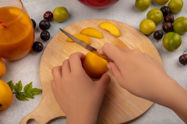 Draufsicht der weiblichen Hände, die gelben Pfirsich auf einem hölzernen Küchenbrett mit Messer mit Pfirsichen mit grünen Kirschpflaumen schneiden, die auf einem weißen Hintergrund lokalisiert werden