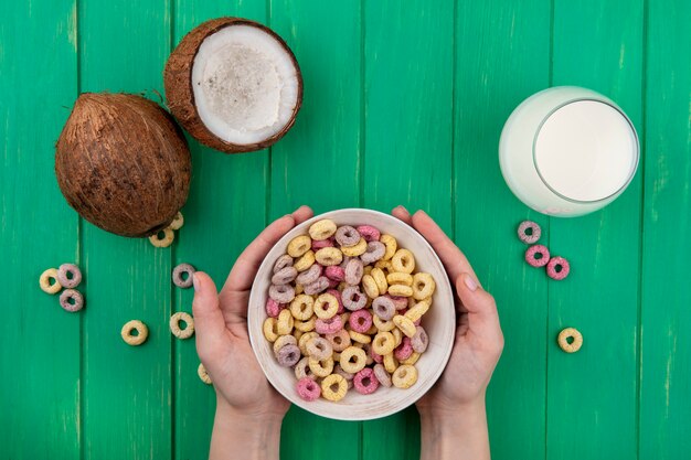 Draufsicht der weiblichen Hände, die Getreide auf weißer Schüssel mit Kokosnüssen und einem Glas Milch auf Grün halten