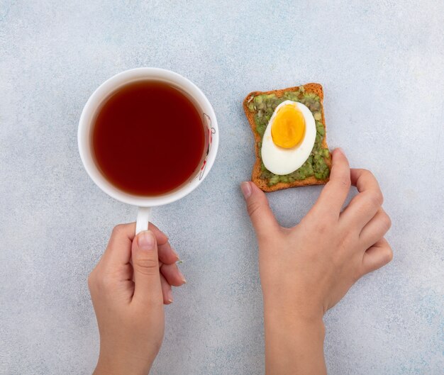 Draufsicht der weiblichen Hände, die in einer Hand Avocado-Scheiben auf Brot und in der anderen Hand Acup von Tee auf Weiß halten