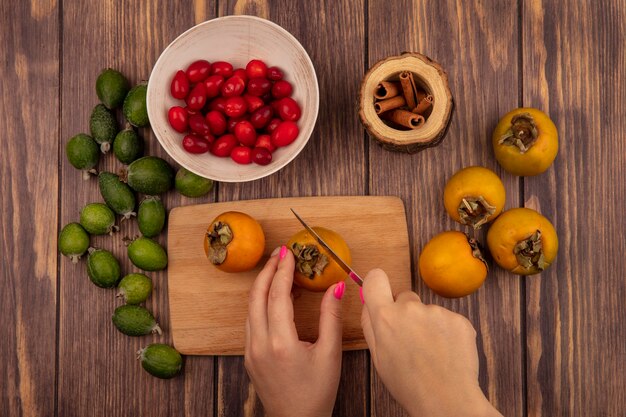 Draufsicht der weiblichen hände, die kakifrucht auf einem hölzernen küchenbrett mit messer mit kornelkirschen auf einer schüssel mit frischer kaki und feijoas schneiden, die auf einem hölzernen hintergrund lokalisiert werden