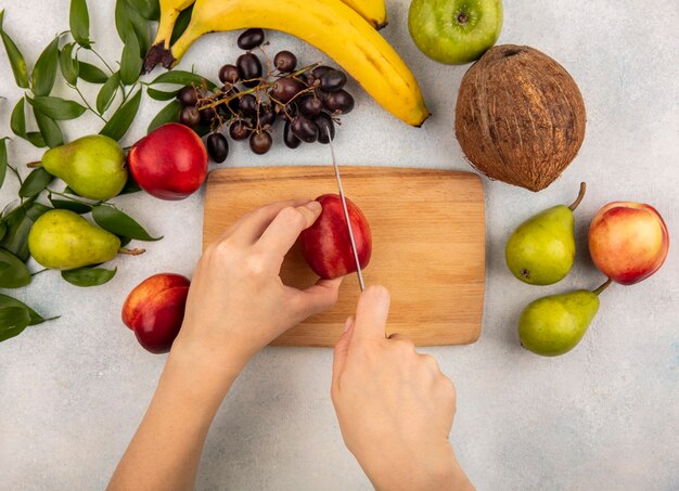 Draufsicht der weiblichen Hände, die Pfirsich mit Messer auf Schneidebrett und Traubenbirnen-Kokosnuss-Bananenapfel mit Blättern auf weißem Hintergrund schneiden