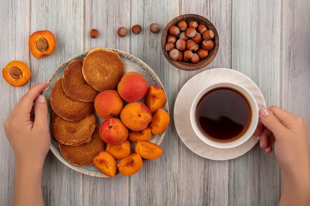 Draufsicht der weiblichen Hände, die Platte Pfannkuchen mit ganzen und geschnittenen Aprikosen und Tasse Tee mit Schüssel Nüssen auf hölzernem Hintergrund halten