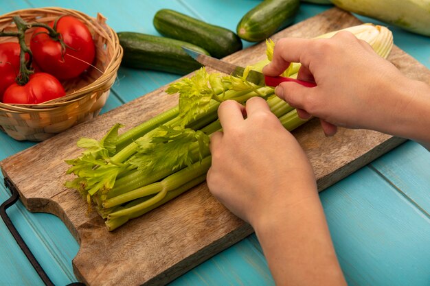 Draufsicht der weiblichen Hände, die Sellerie auf einem hölzernen Küchenbrett mit Messer mit Tomaten auf einem Eimer mit Gurken und Zucchini schneiden, die auf einer blauen hölzernen Oberfläche lokalisiert werden