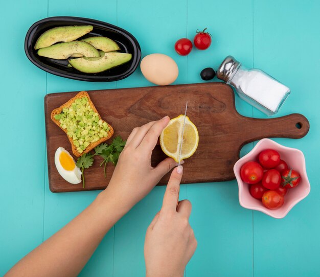 Draufsicht der weiblichen Hände, die Zitrone in Scheiben auf hölzernem Küchenbrett mit Avocado-Scheiben des halben Eiertomaten auf Schüssel auf Blau schneiden