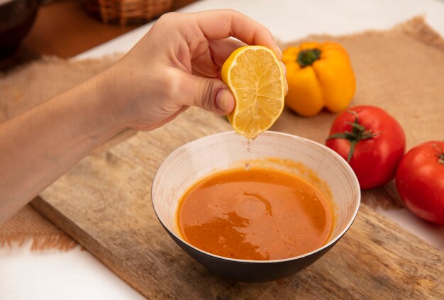 Draufsicht der weiblichen Hand, die eine frische Zitrone in Suppe auf einer Schüssel auf einem hölzernen Küchenbrett auf einem Sackstoff mit bunten Paprikaschoten und Tomaten drückt, die auf einer hölzernen Oberfläche isoliert werden