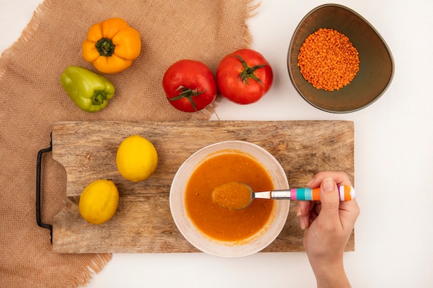 Draufsicht der weiblichen Hand, die einen Löffel mit frischer Linsensuppe auf einer Schüssel auf einem hölzernen Küchenbrett auf einem Sackstoff mit bunten Paprikaschoten und Tomaten lokalisiert auf einer weißen Wand hält