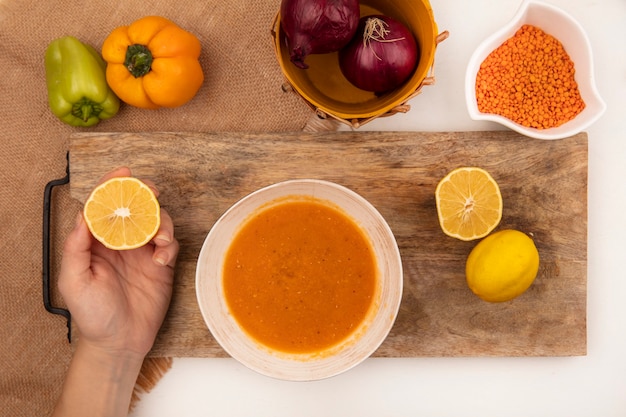 Draufsicht der weiblichen Hand, die frische Zitrone mit Linsensuppe auf einer Schüssel auf einem hölzernen Küchenbrett auf einem Sackstoff mit bunten Paprikaschoten lokalisiert auf einer weißen Oberfläche hält