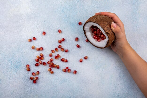 Kostenloses Foto draufsicht der weiblichen hand, die halbe kokosnuss mit roten granatapfelkernen auf weiß hält
