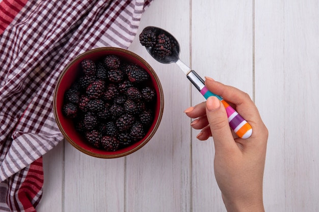 Draufsicht der weiblichen hand hält teelöffel mit einer brombeere mit einem roten karierten handtuch auf einer weißen oberfläche