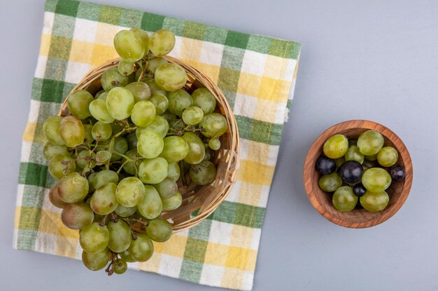 Draufsicht der weißen Traube im Korb auf kariertem Stoff und Traubenbeeren in der Schüssel auf grauem Hintergrund