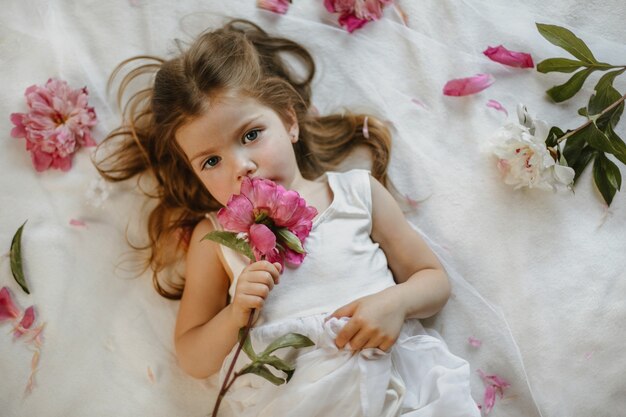 Draufsicht des charmanten aufwachsenden kleinen Mädchens, das eine dunkle rosa Pfingstrose hält, die auf einem weißen Bettlaken liegt, umgeben von frischen Blumen, ernsthaft geradeaus schauend