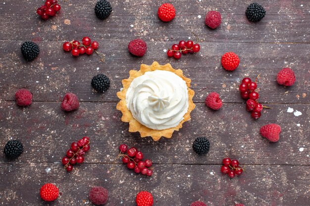 Draufsicht des cremigen köstlichen Kuchens zusammen mit verschiedenen Beeren, die überall auf dem braunen Schreibtisch verteilt werden, Beerenfruchtcreme backen Keks