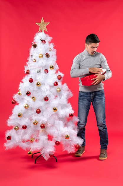 Draufsicht des ernsten gutaussehenden Erwachsenen in einer grauen Bluse, die nahe dem geschmückten weißen Weihnachtsbaum steht