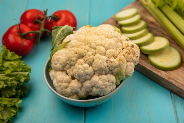 Draufsicht des frischen Blumenkohls auf einer Schüssel mit gehackten Zucchini auf einem hölzernen Küchenbrett mit Sellerie mit Tomaten lokalisiert auf einer blauen Holzwand