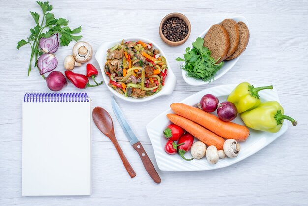 Draufsicht des frischen Gemüsesalats, der mit Fleisch zusammen mit Brotlaib und ganzem Gemüse und Gemüse auf hellem Schreibtisch geschnitten wird, Gemüsesalat