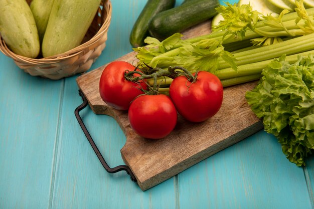 Draufsicht des frischen Selleries auf einem hölzernen Küchenbrett mit Tomatensalat mit Zucchini auf einem Eimer auf einer blauen Holzoberfläche