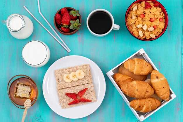 Draufsicht des Frühstücks mit Croissants, Cornflakes, Früchten, Milch und Honig auf blauer Oberfläche horizontal