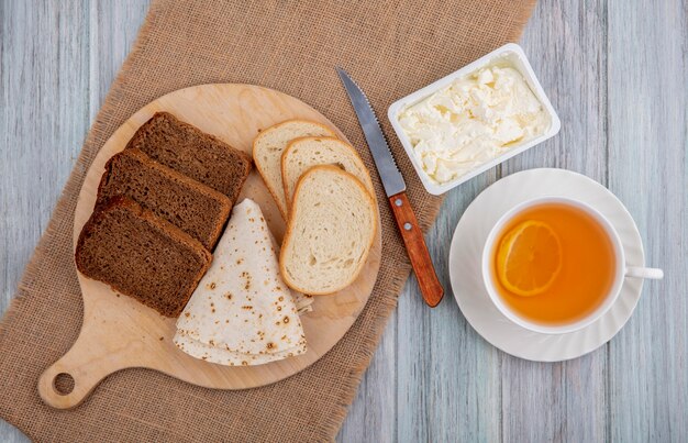 Draufsicht des Frühstückssatzes mit Broten als geschnittenen Roggenweiß und Fladenbrot auf Schneidebrett mit Messer und geronnener Sahne auf Sackleinen und Tasse heißem Wirbel auf hölzernem Hintergrund