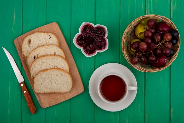 Draufsicht des Frühstückssatzes mit Brotscheiben auf Schneidebrett Himbeermarmelade und Tasse Tee mit Pluot und Traube in Korb und Messer auf grünem Hintergrund